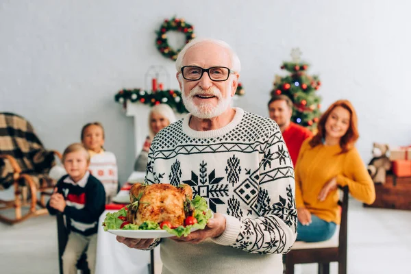 Happy senior man in eyeglasses holding tasty roasted turkey during festive dinner with family — Stock Photo