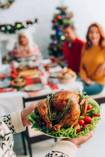 Anciano sosteniendo delicioso pavo asado cerca de la familia sentado en la mesa festiva - foto de stock