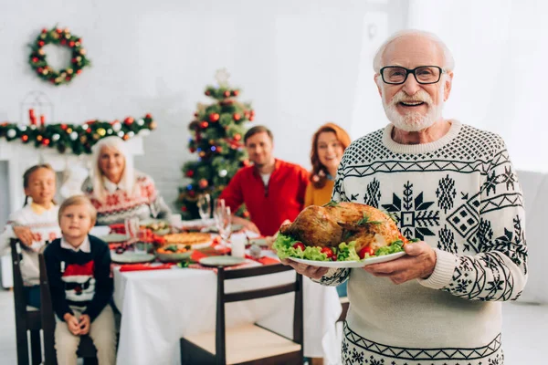 Happy senior man in eyeglasses holding plate with tasty roasted turkey during festive dinner with family — Stock Photo