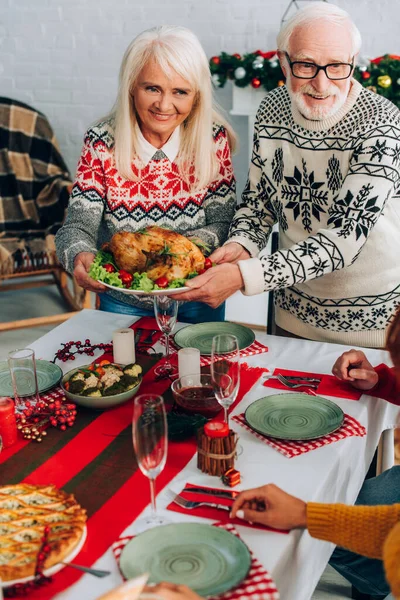 Abuelos sonrientes sirviendo pavo en la mesa festiva cerca de la familia en casa - foto de stock