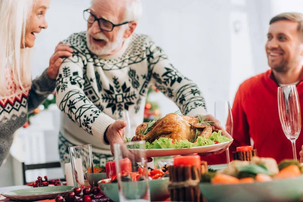 Focus selettivo dell'uomo anziano che serve tacchino sul tavolo festivo in piedi vicino alla moglie — Foto stock