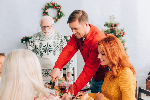 Улыбающийся мужчина режет индейку на праздничном столе с ужином рядом с семьей дома — стоковое фото