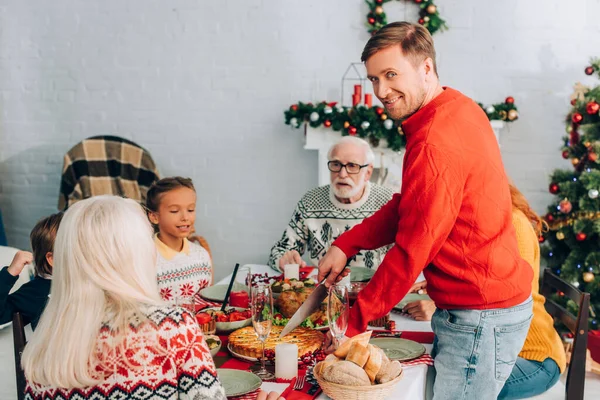 Uomo sorridente guardando la fotocamera mentre taglia torta sul tavolo festivo vicino alla famiglia — Foto stock