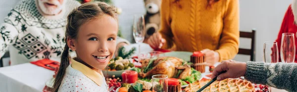 Récolte panoramique de fille regardant la caméra, assis à la table festive près de la famille — Photo de stock