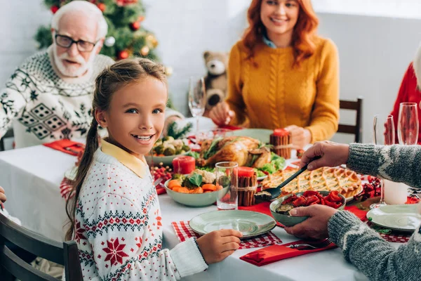 Foco seletivo da menina olhando para a câmera, sentado na mesa festiva perto da família — Fotografia de Stock
