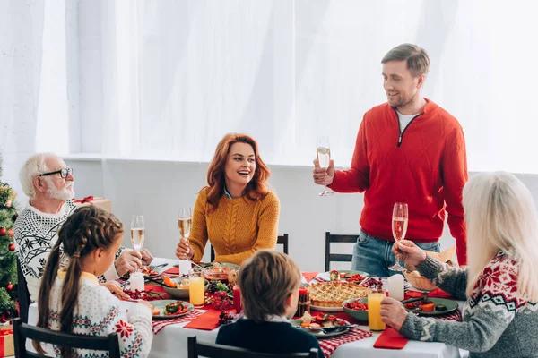 Homem sorridente com taça de champanhe dizendo discurso perto da família na mesa festiva em casa — Fotografia de Stock