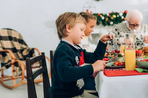 Selektiver Fokus des kleinen Jungen, der mit Messer und Gabel isst, am Tisch sitzt — Stockfoto