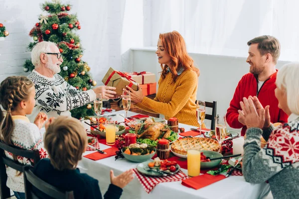 Femme rousse saluant homme âgé avec boîte cadeau, assis à table avec la famille — Photo de stock