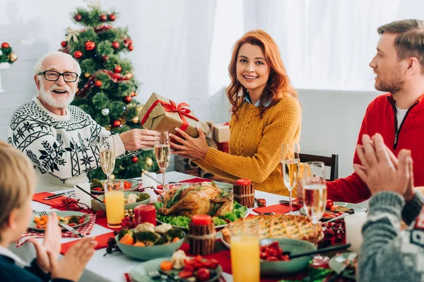 Femme et homme âgé tenant boîte cadeau tout en étant assis avec la famille à la table de fête — Photo de stock