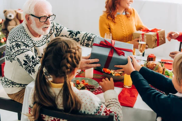 Nonno saluto nipote con scatola regalo vicino alla famiglia seduta a tavola festiva — Foto stock