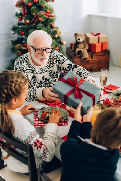 Grandfather giving present to grandson while sitting with family near at table — Stock Photo