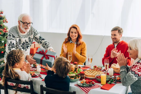 Smiling grandfather giving present to grandson near family applauding at home — Stock Photo