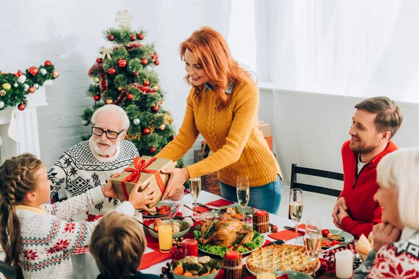 Madre saludo hija con presente, de pie cerca de la familia en la mesa festiva - foto de stock