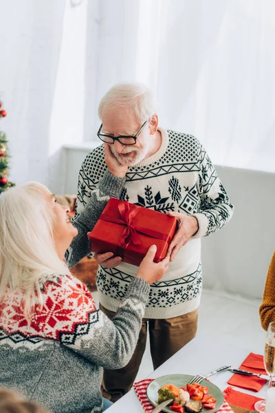 Senior woman holding hand on husband cheek while taking gift box at home — Stock Photo