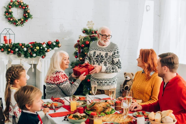 Felice coppia anziana in possesso di scatola regalo vicino alla famiglia seduta al tavolo festivo — Foto stock