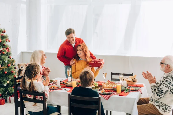 Wife holding gift box from husband laughing and sitting near family — Stock Photo