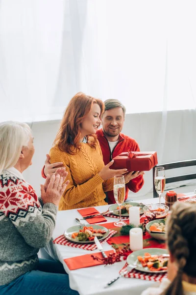 Marito abbracciando moglie guardando scatola regalo, seduto con la famiglia al tavolo festivo — Foto stock