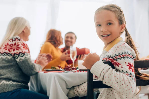 Selektiver Fokus eines lächelnden Mädchens, das in die Kamera blickt, mit der Familie am Tisch sitzt — Stockfoto