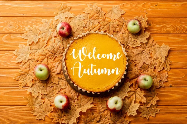 Vista superior de torta de abóbora de ação de graças com maçãs em folhas perto de lettering outono bem-vindo no fundo de madeira — Fotografia de Stock