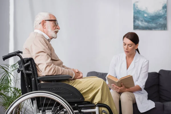 Enfermera geriátrica leyendo libro a hombre discapacitado envejecido sobre fondo borroso - foto de stock
