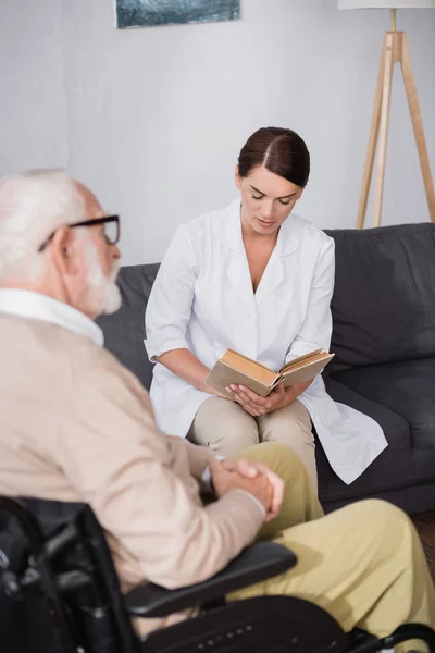 Social worker reading book to aged man on blurred foreground — Stock Photo