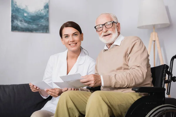 Sonriente trabajador social y anciano hombre mirando a la cámara mientras sostiene fotos - foto de stock