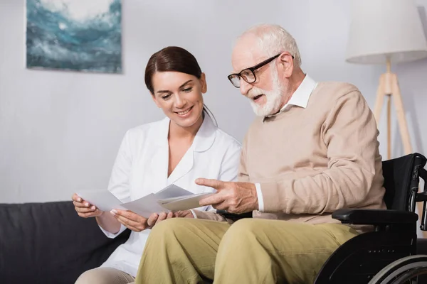 Cheerful elderly man and smiling social worker looking at photos at home — Stock Photo