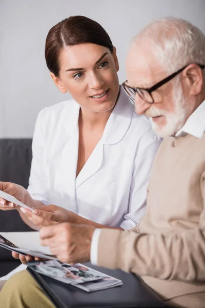 Brunette social worker looking at aged mad while holding book on blurred foreground — Stock Photo