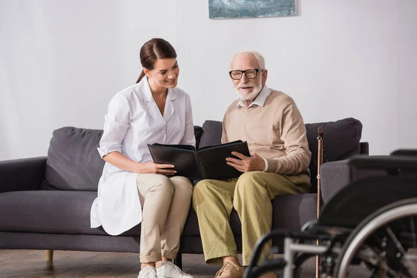 Lächelnder älterer Mann blättert im Fotoalbum, während er mit Sozialarbeiterin auf dem Sofa sitzt, verschwommener Vordergrund — Stockfoto