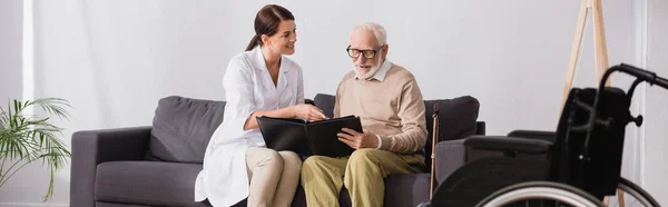 Altenpfleger und älterer Mann sehen sich Fotos an, während sie auf dem Sofa sitzen — Stockfoto