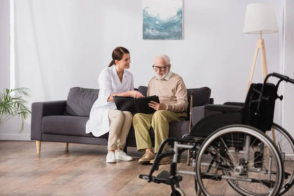 Enfermeiro geriátrico e homem idoso navegando álbum de fotos perto cadeira de rodas em primeiro plano borrado — Fotografia de Stock