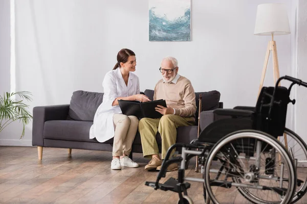 Infirmière gériatrique et homme âgé parcourant l'album photo près du fauteuil roulant au premier plan flou — Photo de stock