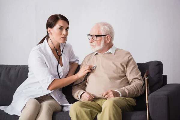 Social worker checking health of aged patient with stethoscope — Stock Photo