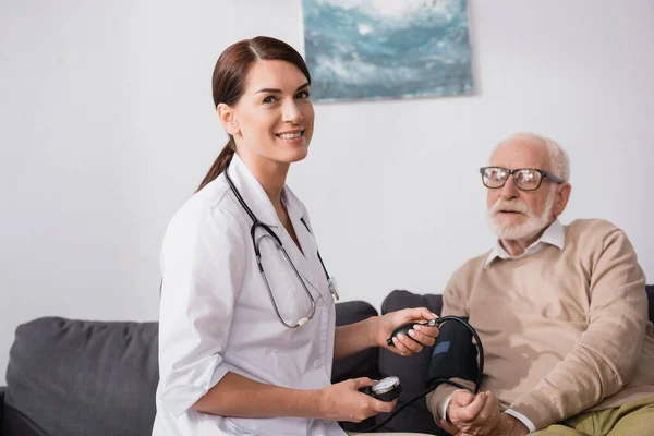 Smiling social worker looking at camera while checking health of patient with stethoscope — Stock Photo