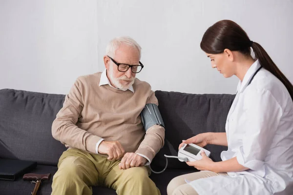 Enfermeira geriátrica morena examinando homem idoso com tonômetro em casa — Fotografia de Stock
