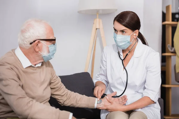Социальный работник в медицинской маске осматривает пожилого человека со стетоскопом дома — стоковое фото