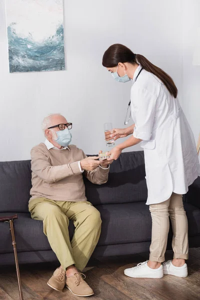 Sozialarbeiterin gibt älteren Mann in medizinischer Maske Wasser und Tabletten — Stockfoto
