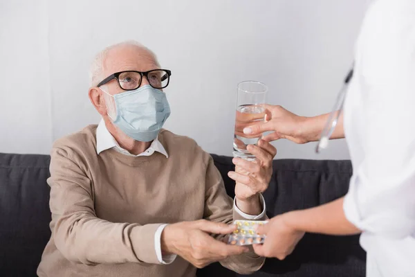 Älterer Mann in medizinischer Maske nimmt Glas Wasser und Tabletten von Altenpflegerin auf verschwommenem Vordergrund — Stockfoto