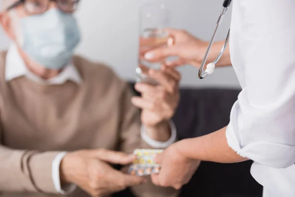Cropped view of social worker giving medicines and water to elderly man on blurred background — Stock Photo