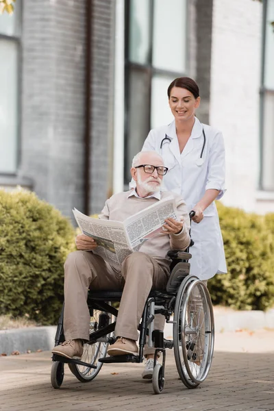 Smiling geriatric nurse walking with aged disable man holding newspaper in wheelchair outdoors — Stock Photo