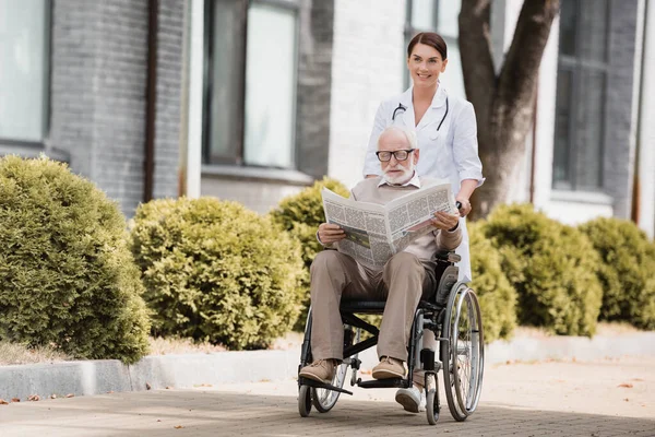 Cheerful social worker walking with disabled elderly man reading newspaper in wheelchair — Stock Photo