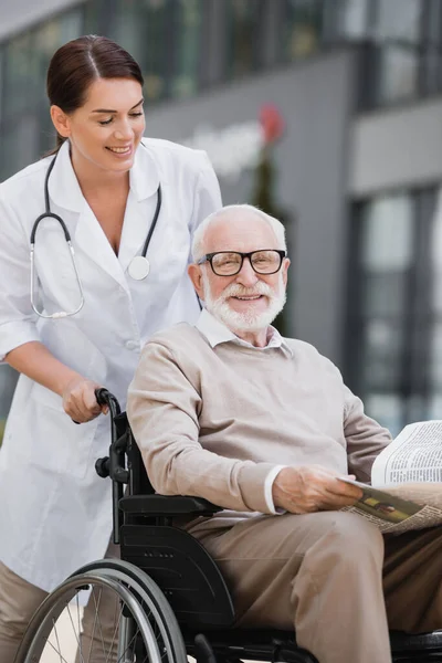 Smiling social worker walking with cheerful aged man in wheelchair holding newspaper outdoors — Stock Photo