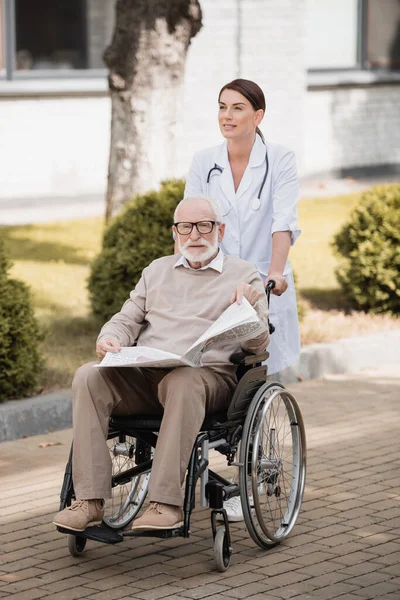 Sozialarbeiterin geht mit gehbehindertem Mann mit Zeitung im Rollstuhl — Stockfoto