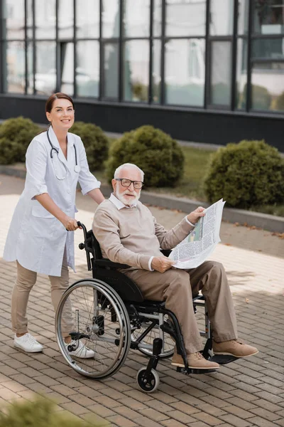 Cheerful social worker looking at camera while walking with handicapped aged man with newspaper outdoors — Stock Photo