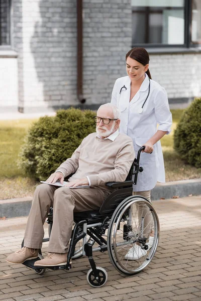 Brunette social worker walking with aged handicapped man in wheelchair outdoors — Stock Photo