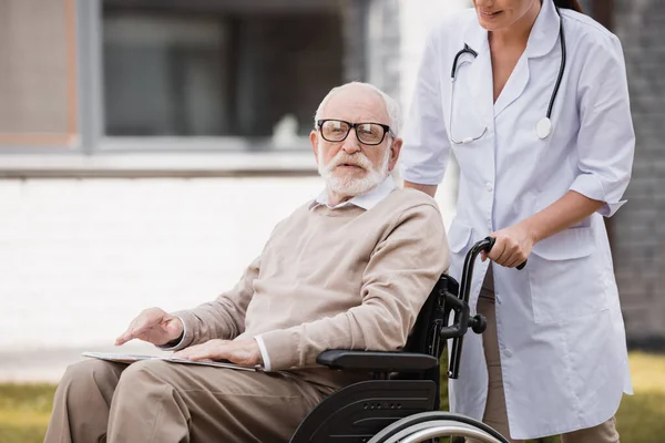 Aged handicapped man in wheelchair looking at camera while walking outdoors with geriatric nurse — Stock Photo