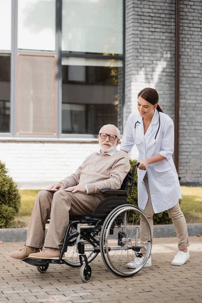 Cheerful social worker walking with elderly handicapped man in wheelchair outdoors — Stock Photo