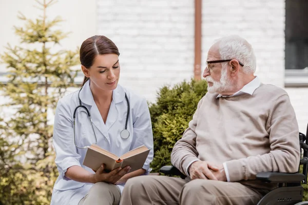 Geriatric nurse reading book for disabled man in wheelchair outdoors — Stock Photo