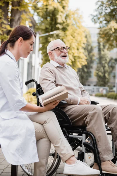 Social worker reading book to cheerful disabled man in wheelchair outdoors — Stock Photo