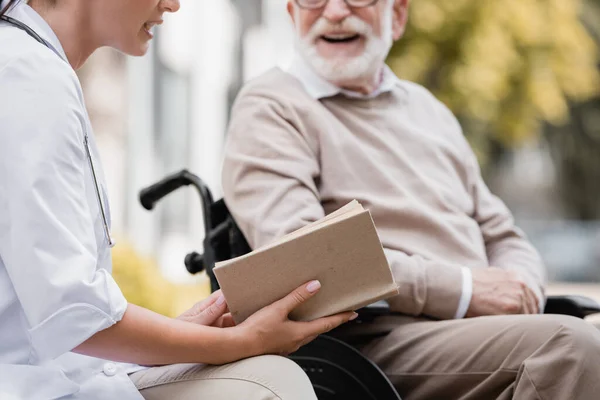 Cropped view of geriatric nurse reading book to aged man on blurred background — Stock Photo